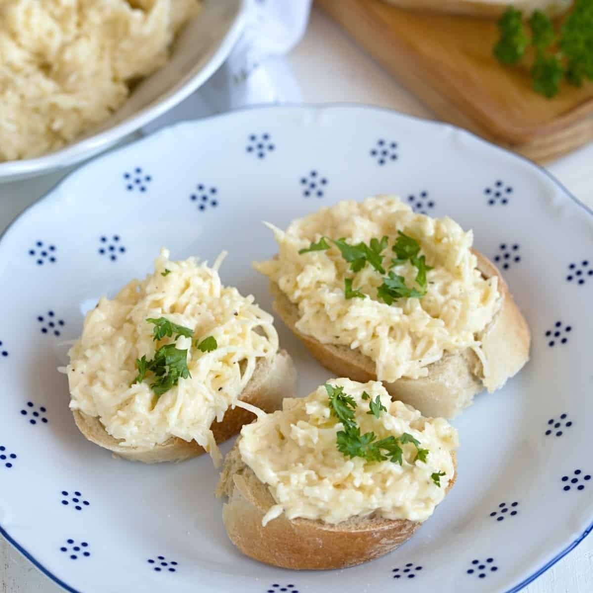 Brötchen-Scheiben bestrichen mit Knoblauch-Käse-Aufstrich, angerichtet an weissem Teller. 